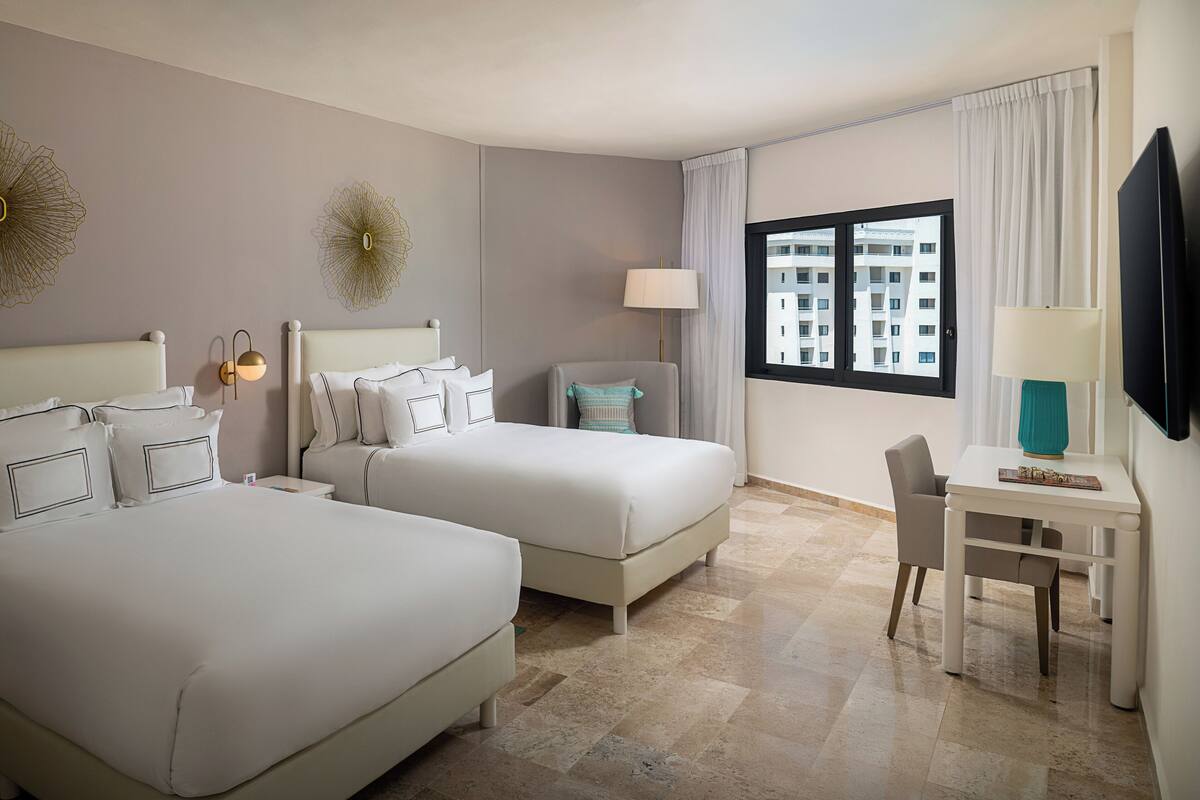 A hotel room with two beds, a desk, a chair, a TV, and a window with a city view.