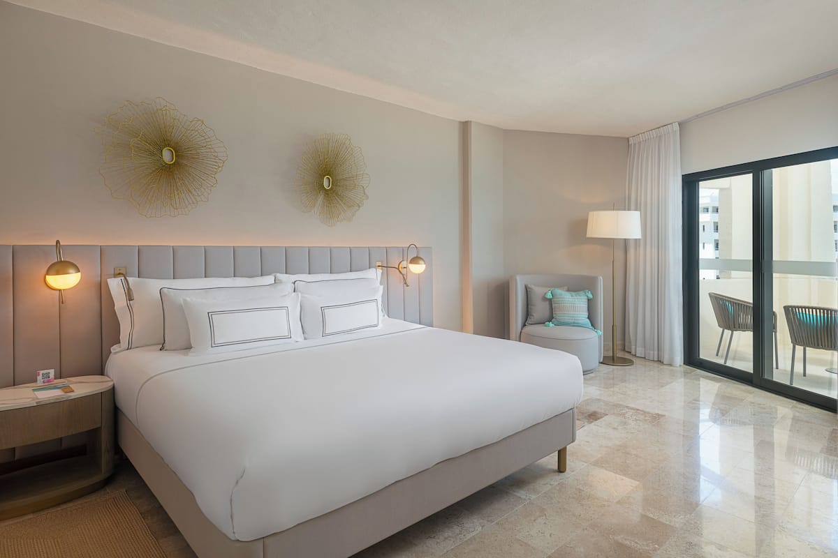 A hotel room with a large bed, two wall sconces, a round side table, a chair, and a view through a large window.