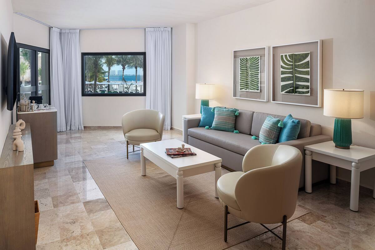 A modern living room with a sofa, two armchairs, a coffee table, and a TV on a stand. There are two side tables with lamps, a large window with a view of palm trees and the sea, and a small desk with a mirror and decorative items.