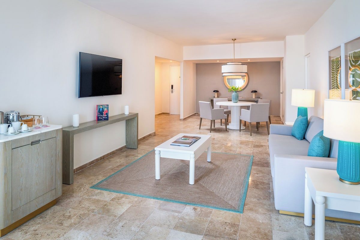 A modern living room with a flat-screen TV, a dining area with a round table and chairs, a sofa with cushions, and a coffee table.