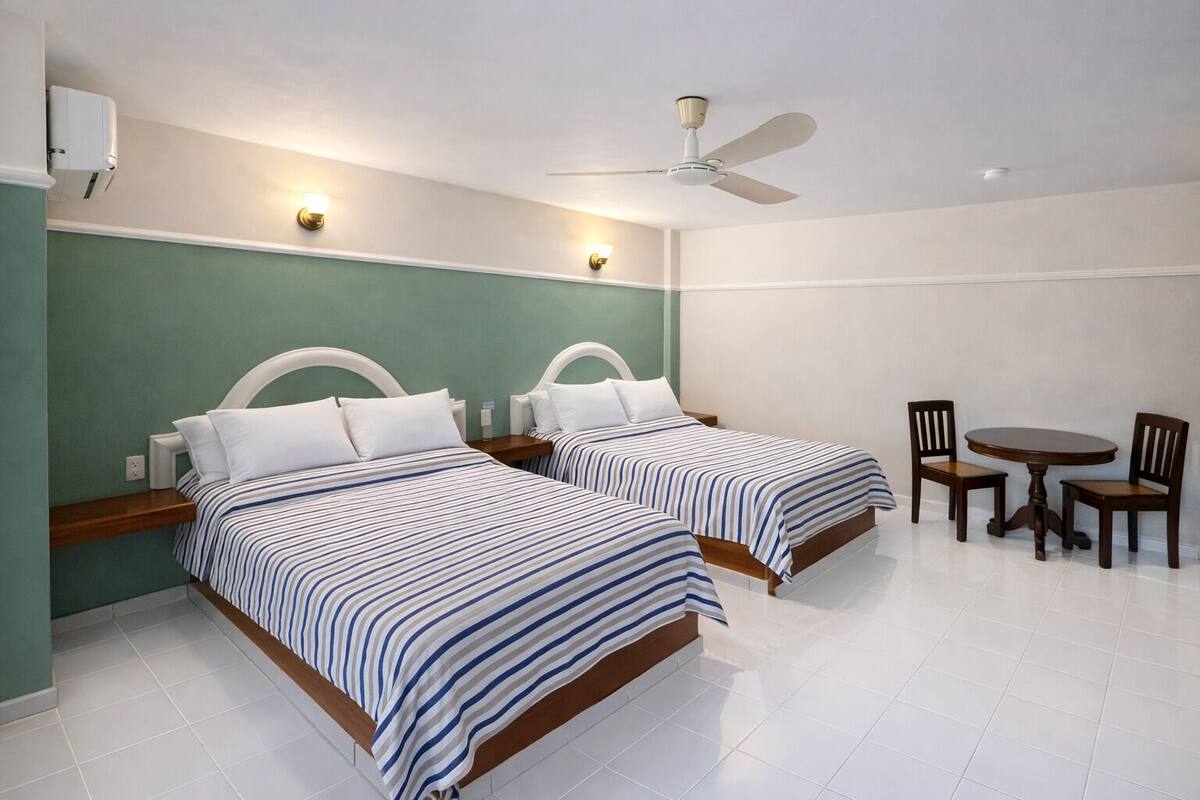 A hotel room with two beds, a ceiling fan, a small round table with chairs, and a wall-mounted air conditioning unit.