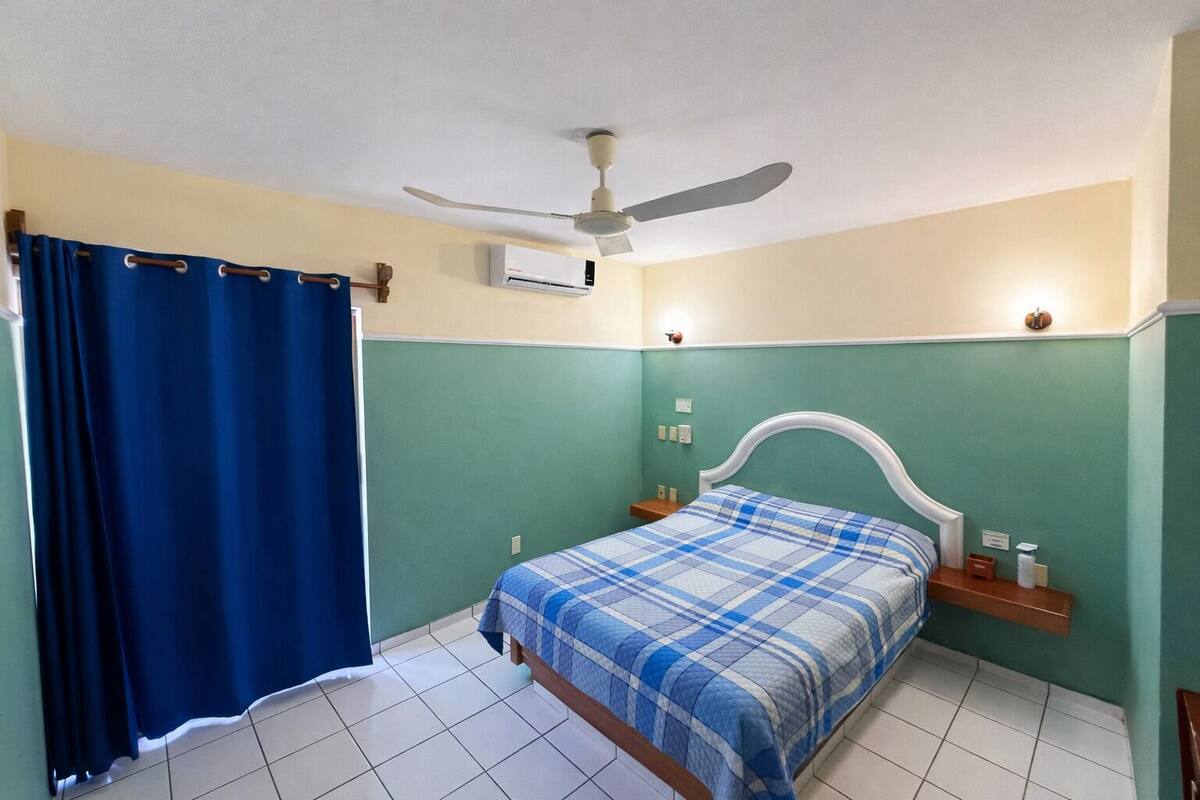 A bedroom with a bed, a ceiling fan, an air conditioning unit, a nightstand, and a blue curtain.