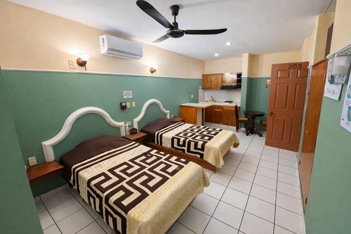 A hotel room with two beds, a desk, a chair, a ceiling fan, an air conditioning unit, and a door with the number 203.