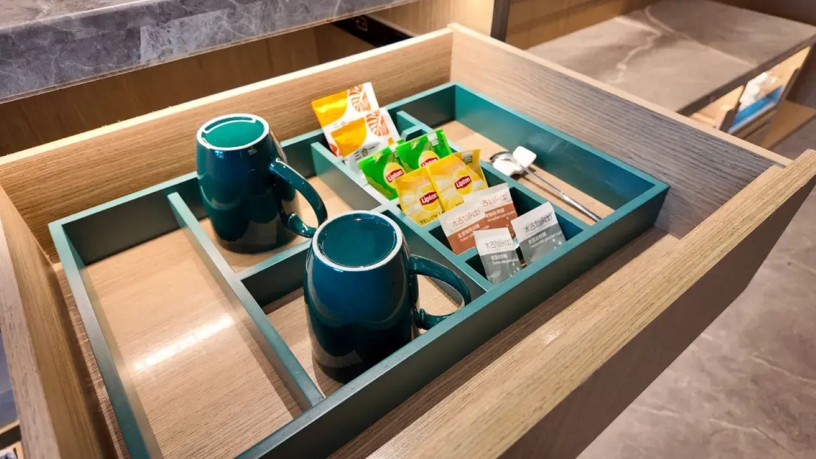 Un cassetto suddiviso in scomparti con una tazza blu, una tazza verde e pacchetti di tè Lipton.