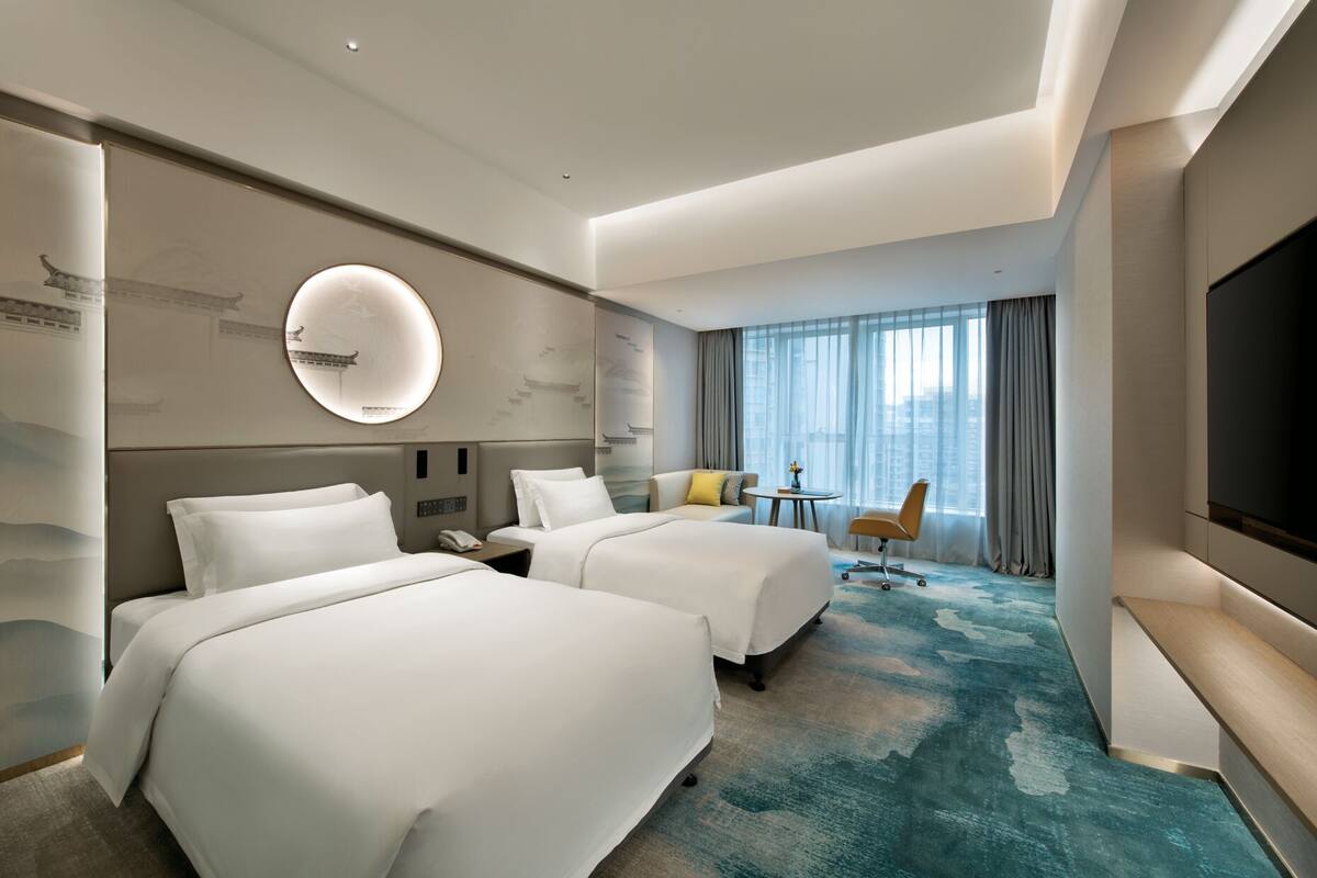 Une chambre d’hôtel avec deux lits, un miroir rond, un bureau, une chaise et une grande télévision.