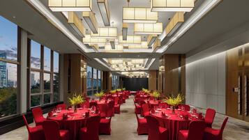 A spacious conference room with multiple tables set for dining, large windows offering a city view, and modern ceiling lighting.