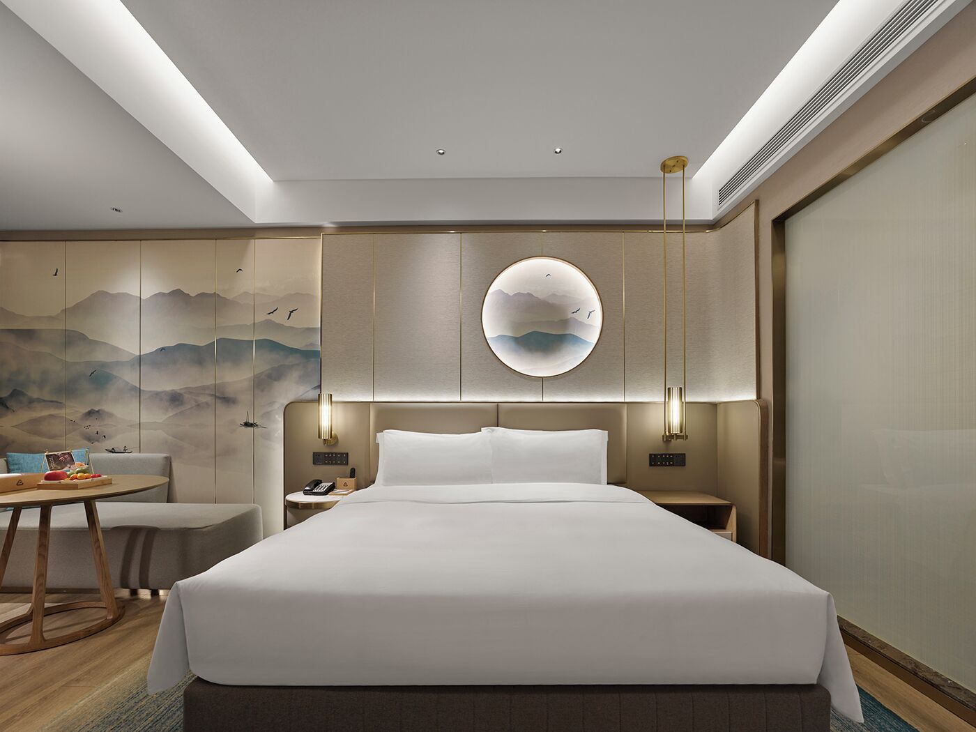 A modern hotel room with a large bed, a round wall mirror, a desk with a chair, and a wall mural.