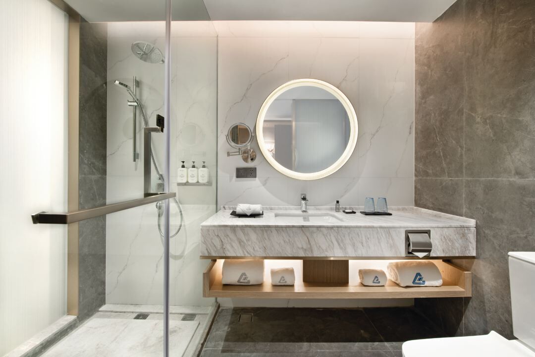 Une salle de bain moderne dotée d’une douche à parois de verre, d’un meuble-lavabo en marbre, d’un miroir rond et de toilettes avec un porte-serviettes.