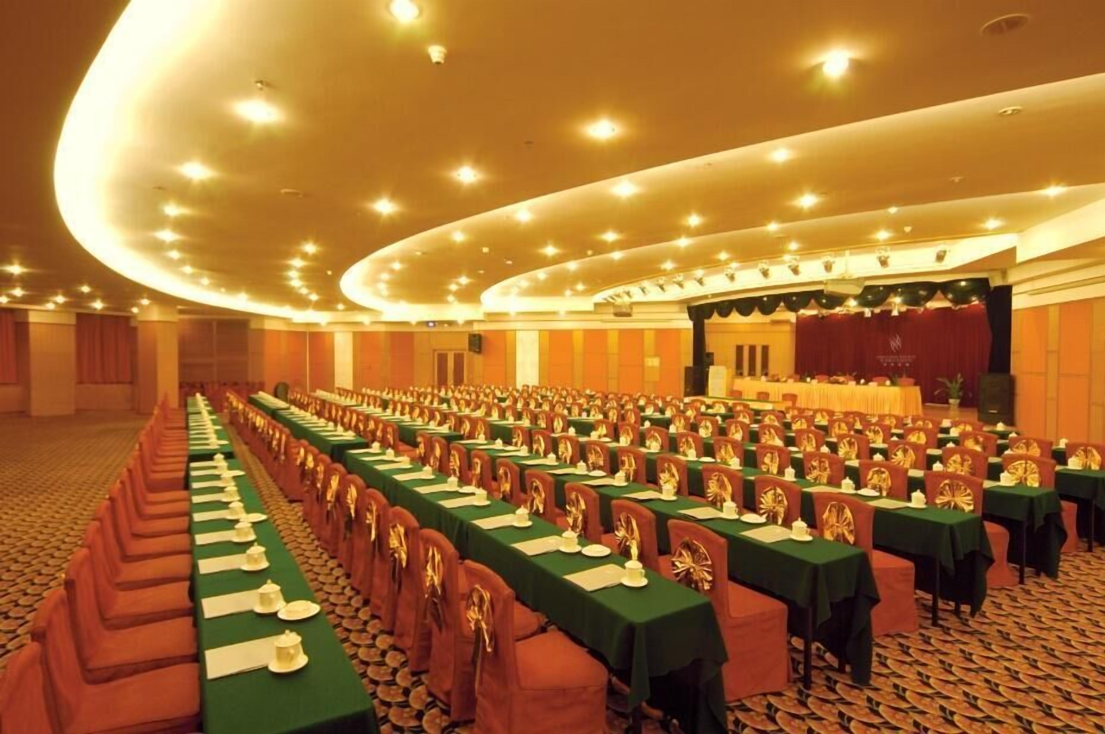 A large conference room with rows of tables and chairs, a stage with a curtain, and a carpeted floor.