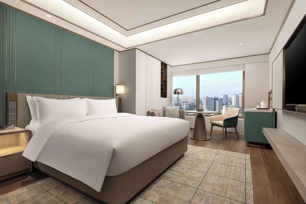 A modern hotel room with a large bed, a desk, a chair, and a view of the city skyline.