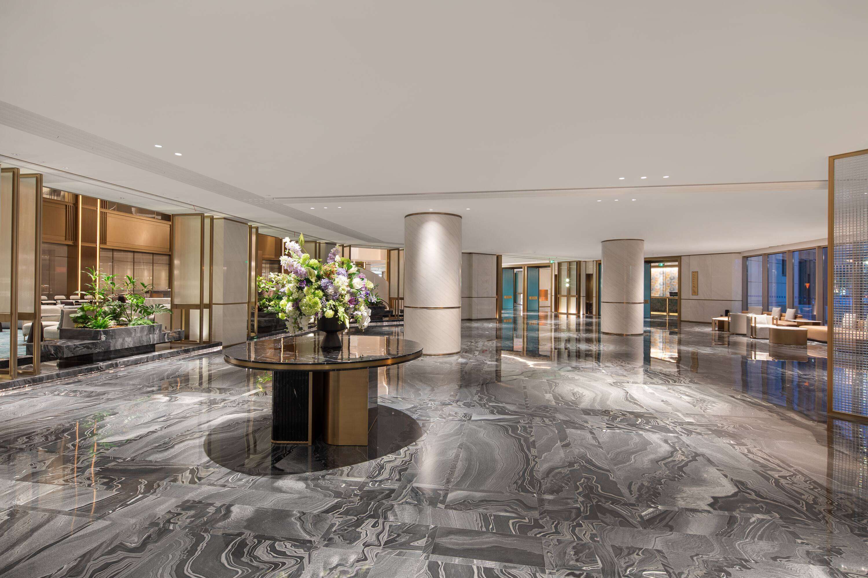 Um hall amplo com piso de mármore, uma mesa redonda central com arranjo floral e áreas de estar com decoração moderna.