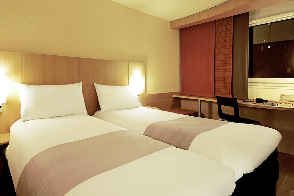Habitación de hotel con dos camas, un escritorio, una silla y una ventana con cortinas.