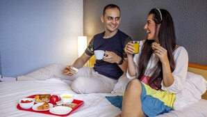 A couple sitting on a bed with breakfast items including toast, fruit, and a drink. - ibis Criciúma (Criciuma)