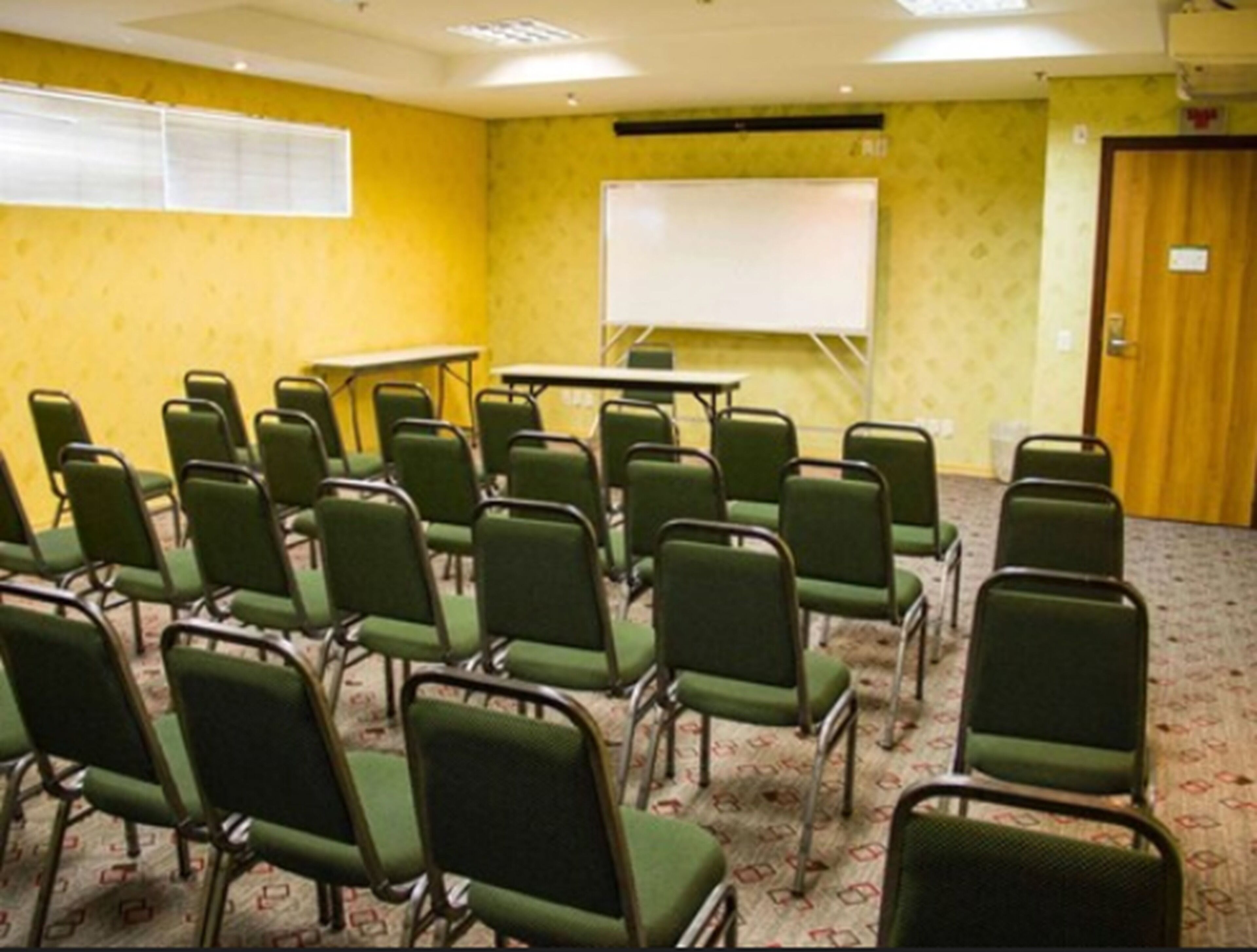 Uma sala de conferências com cadeiras verdes dispostas em fileiras, uma lousa e uma mesa.