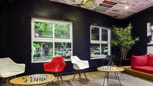 A modern lounge area with a chessboard on a round table, three chairs, a red sofa, and large windows with a view of parked cars. - ibis Criciúma (Criciuma)