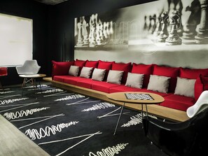 Un salon moderne avec un canapé rouge, des fauteuils blancs, une table d’échecs et un écran de projection.