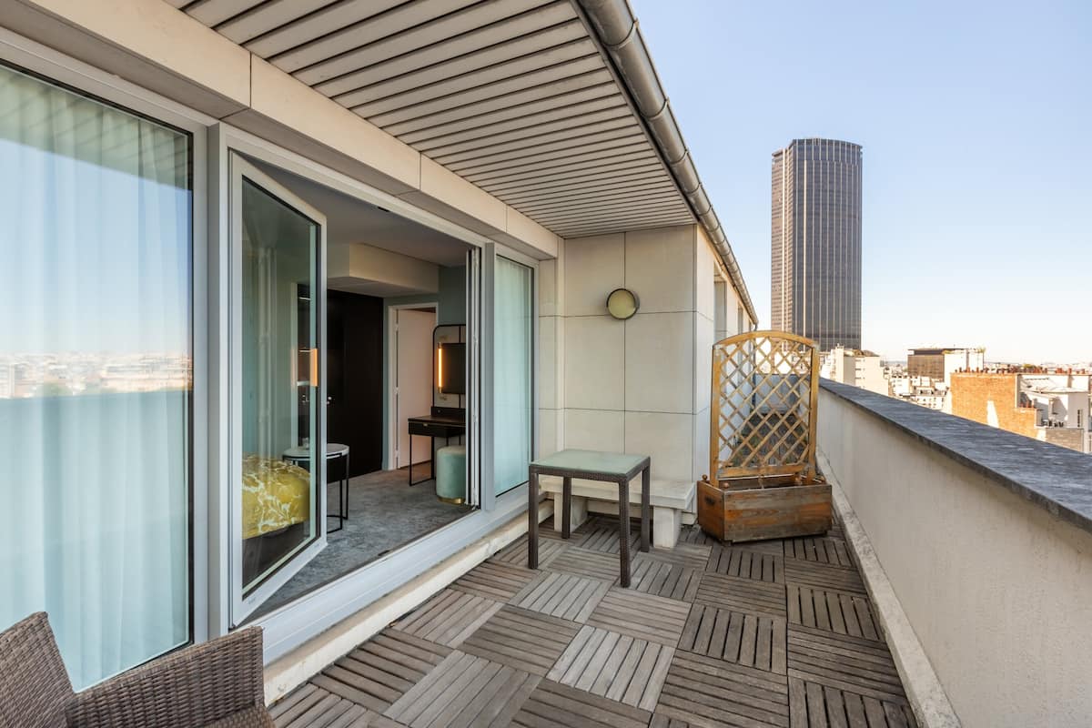 A balcony with a view of the city, a wooden floor, a small table, and a wicker chair.