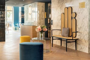 A reception area with a marble wall, a blue chair, a yellow ottoman, and a vase with flowers.
