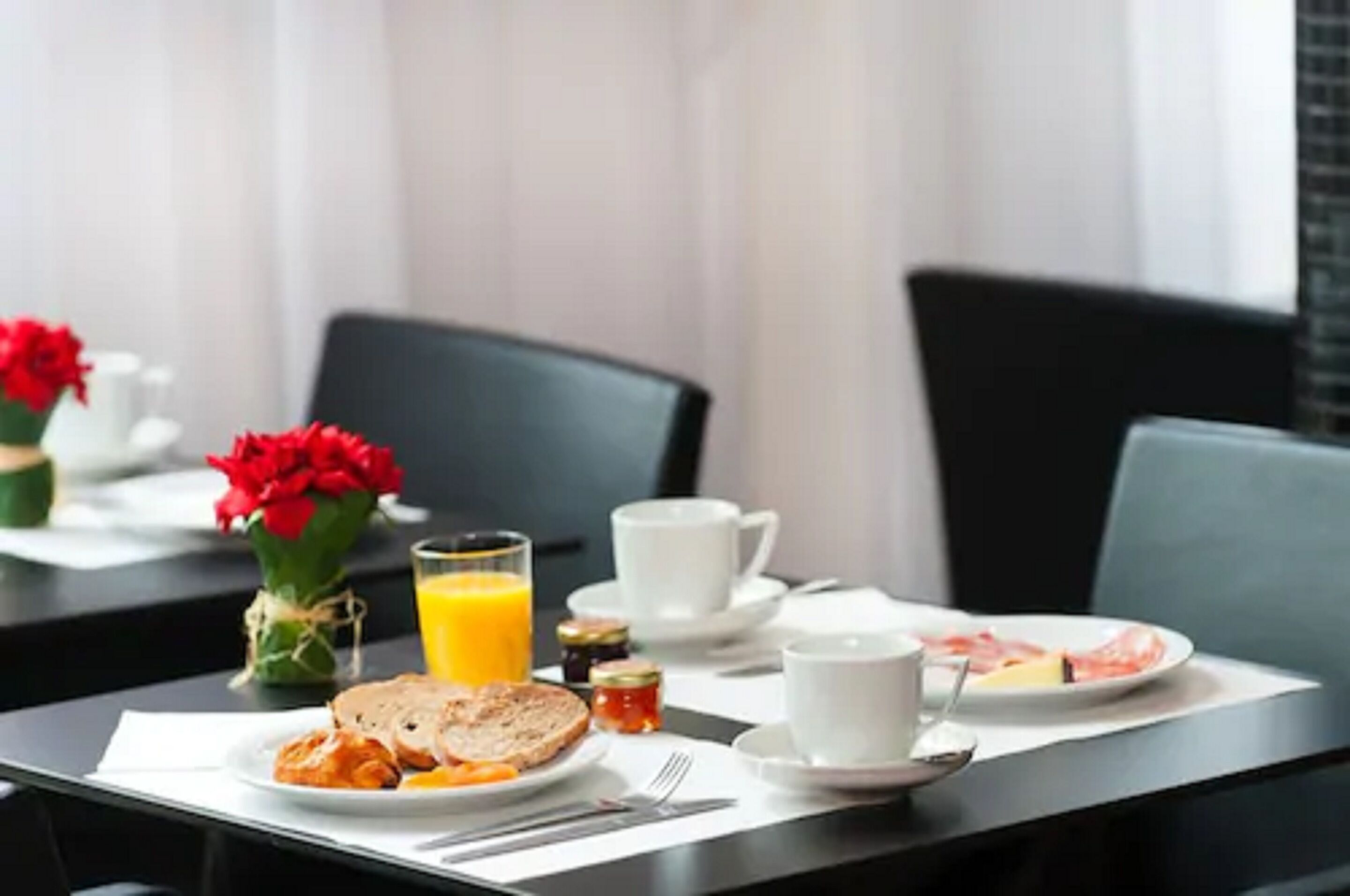 A breakfast table set with plates, toast, jam, a glass of orange juice, and a vase of red flowers.