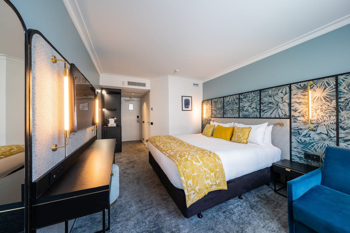 A hotel room with a large bed, a desk, a chair, a mirror, and a wall with a floral pattern.