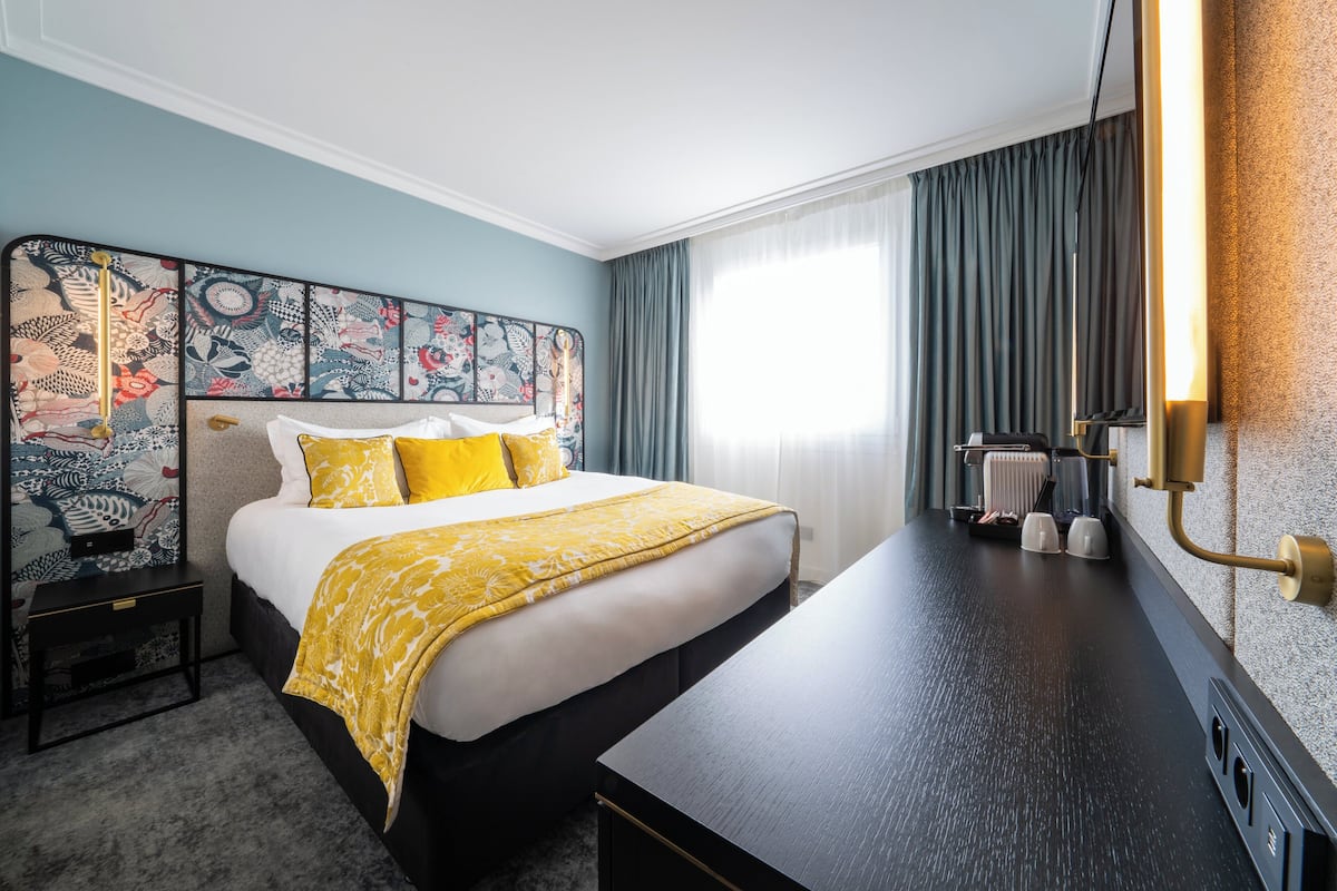 A hotel room with a bed, bedside table, and a desk with a lamp.