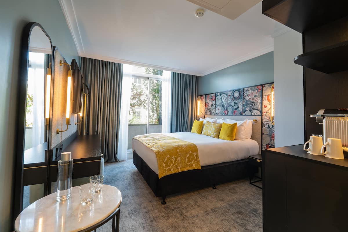 A hotel room with a bed, a desk, a chair, a table with glasses, a mirror, and a window with curtains.