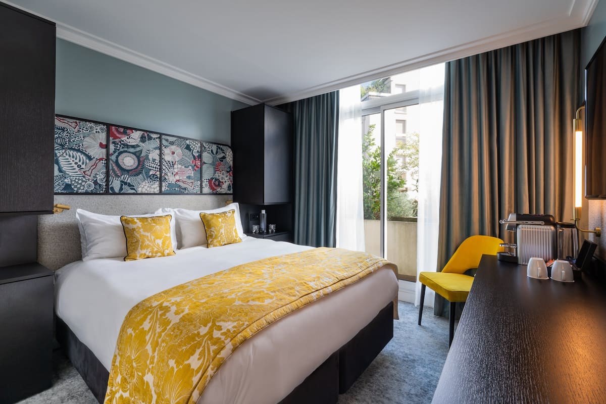 A hotel room with a large bed, a yellow chair, a desk with a coffee machine, and a view of the outdoors through a sliding door.