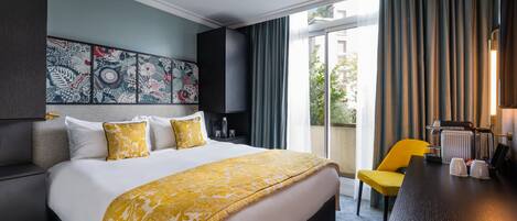 A hotel room with a large bed, a yellow chair, a desk with a coffee machine, and a view of the outdoors through a sliding door.