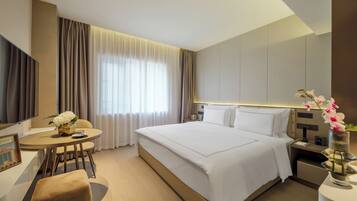 A modern hotel room with a large bed, a desk with a chair, a small table, and a bedside table with a vase of flowers.