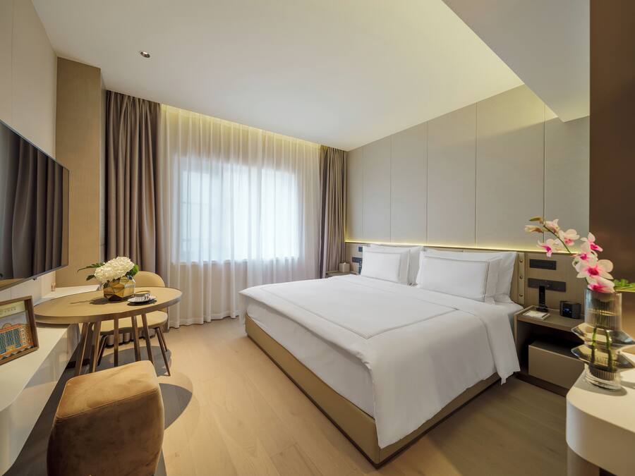 A modern hotel room with a large bed, a desk with a chair, a small table, and a bedside table with a vase of flowers.