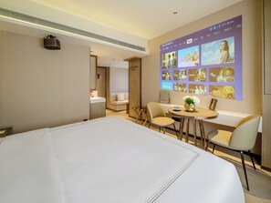 Una habitación de hotel con una cama grande, un escritorio con sillas, una pantalla de televisión mostrando varios programas y un proyector montado en la pared.