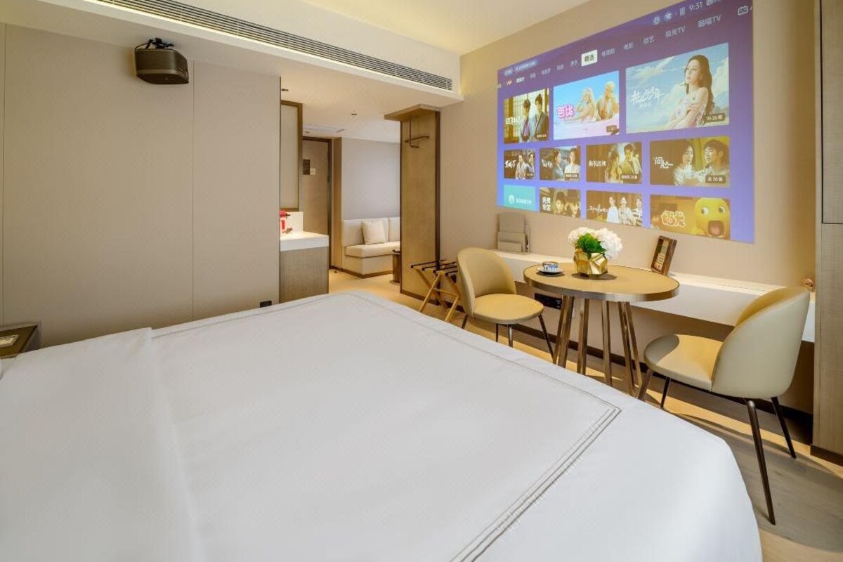 A hotel room with a large bed, a desk with chairs, a small table with a vase, and a wall-mounted TV displaying movie posters.