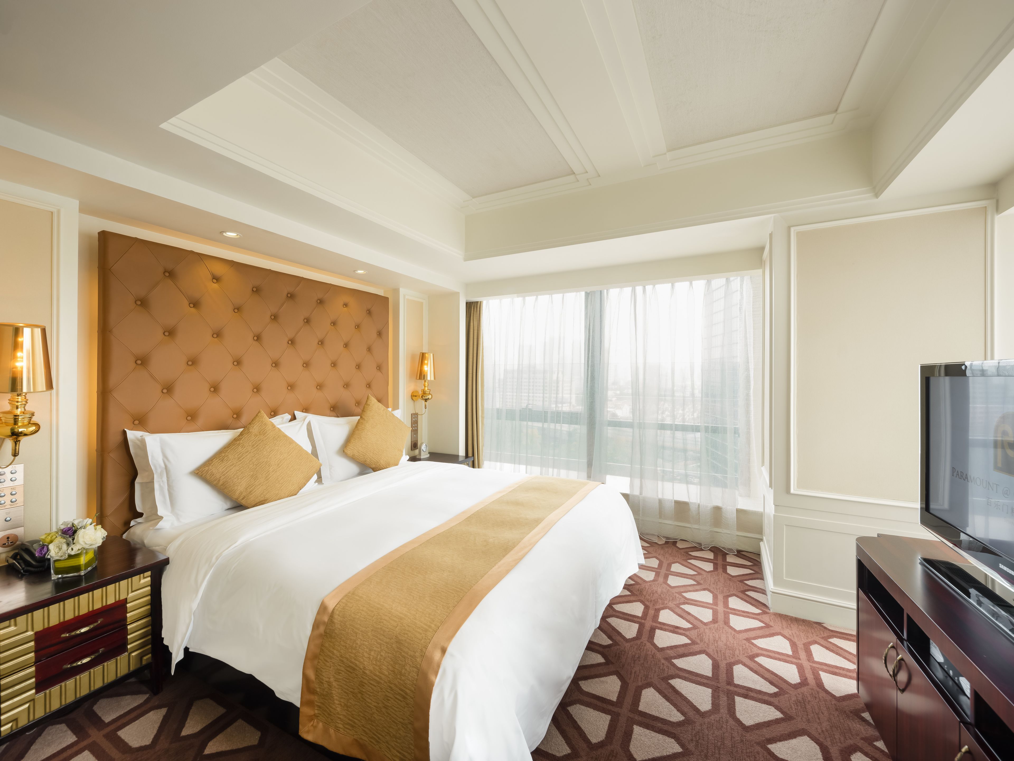 Quarto de hotel com uma cama grande, televisão, mesa de cabeceira e janela com cortinas finas.