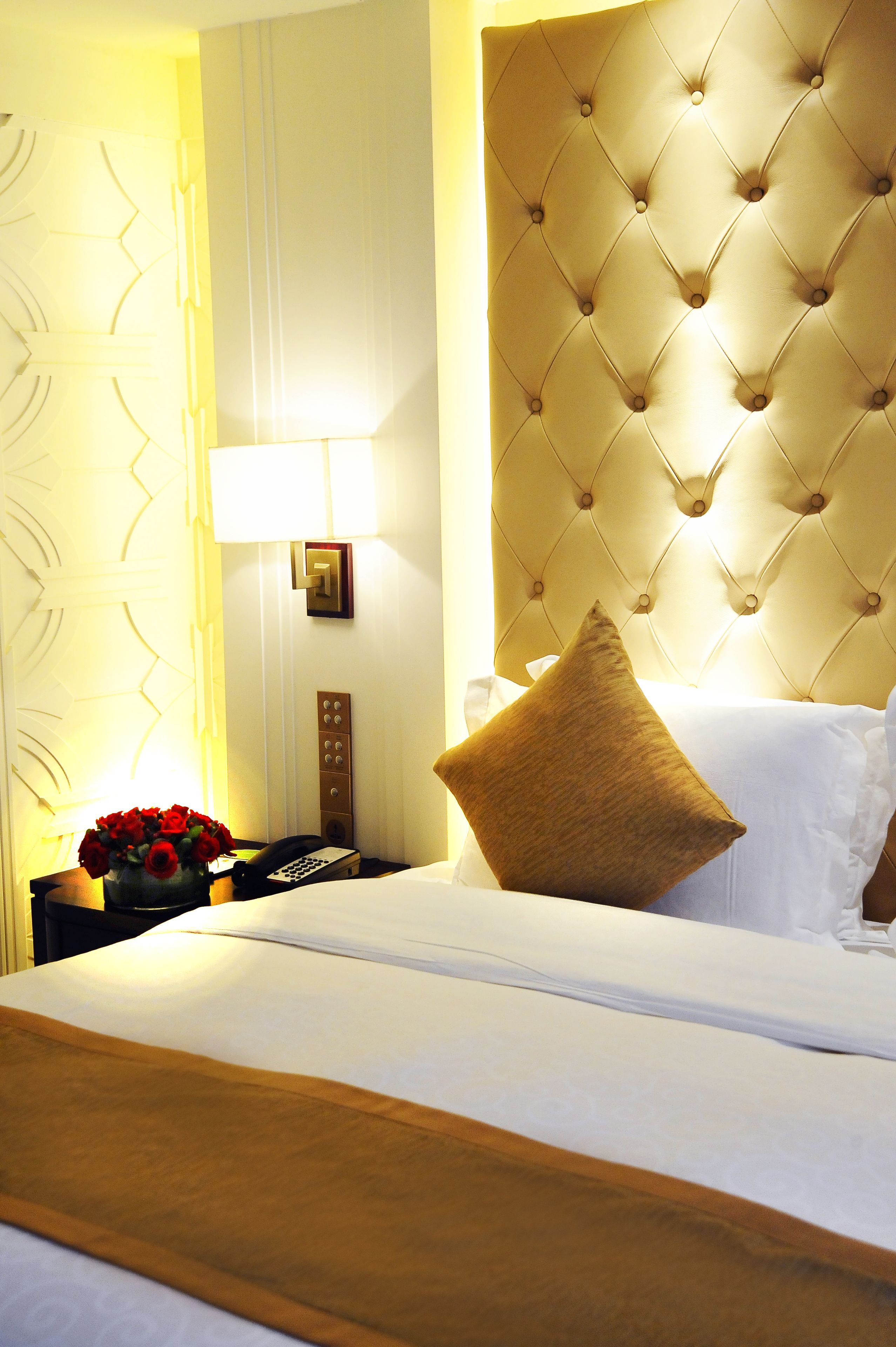 Una habitación de hotel con cabecera capitonada, una mesita de noche con lámpara, un jarrón de rosas rojas y un teléfono.