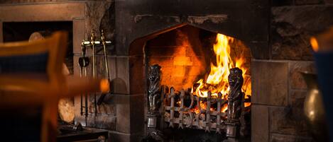 A stone fireplace with a roaring fire, ornate metal grates, and a chimney.