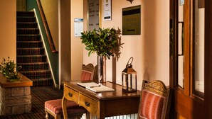 Un hall d’hôtel avec un comptoir d’accueil en bois, une plante en pot, un panneau indiquant les plats proposés au bar, un escalier et un tableau de menu fixé au mur.