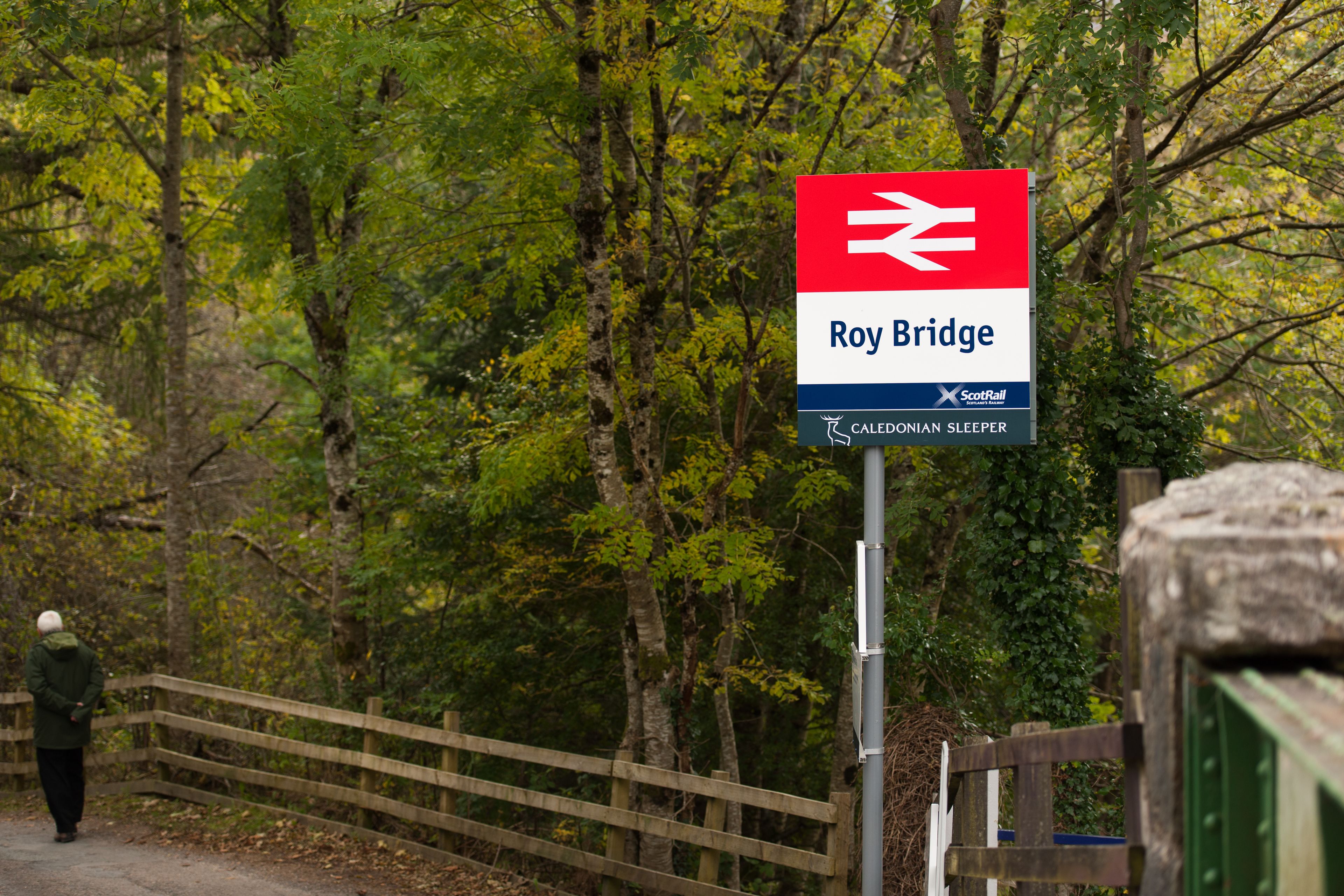 Bordje voor Roy Bridge treinstation, met een houten hek en bomen op de achtergrond.