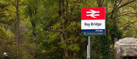 Bordje voor Roy Bridge treinstation, met een houten hek en bomen op de achtergrond.