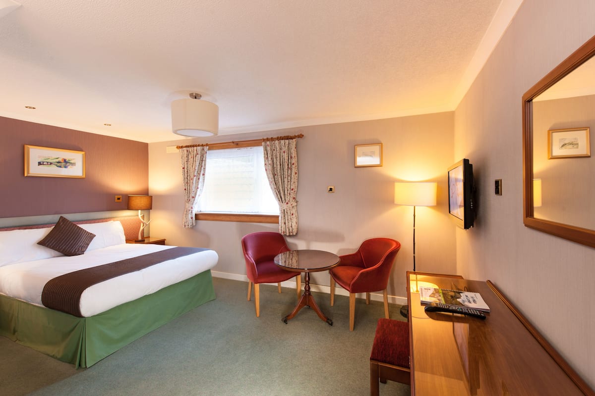 A hotel room with a bed, two red chairs, a small table, a TV, and a window with curtains.