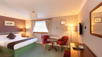 A hotel room with a bed, two red chairs, a small table, a TV, and a window with curtains.