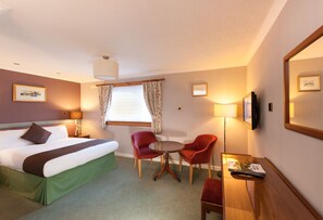 A hotel room with a bed, two red chairs, a small table, a TV, and a window with curtains.