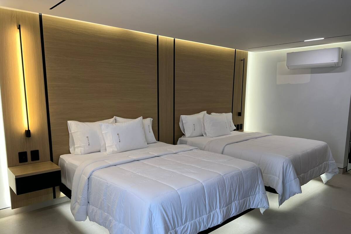 A modern hotel room with two beds, a wooden headboard, a bedside table, and a wall-mounted air conditioner.