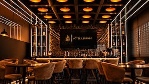 Zona de bar con diseño moderno, con una gran pantalla de televisión mostrando el nombre del hotel, varios arreglos de asientos y una variedad de botellas en estantes.