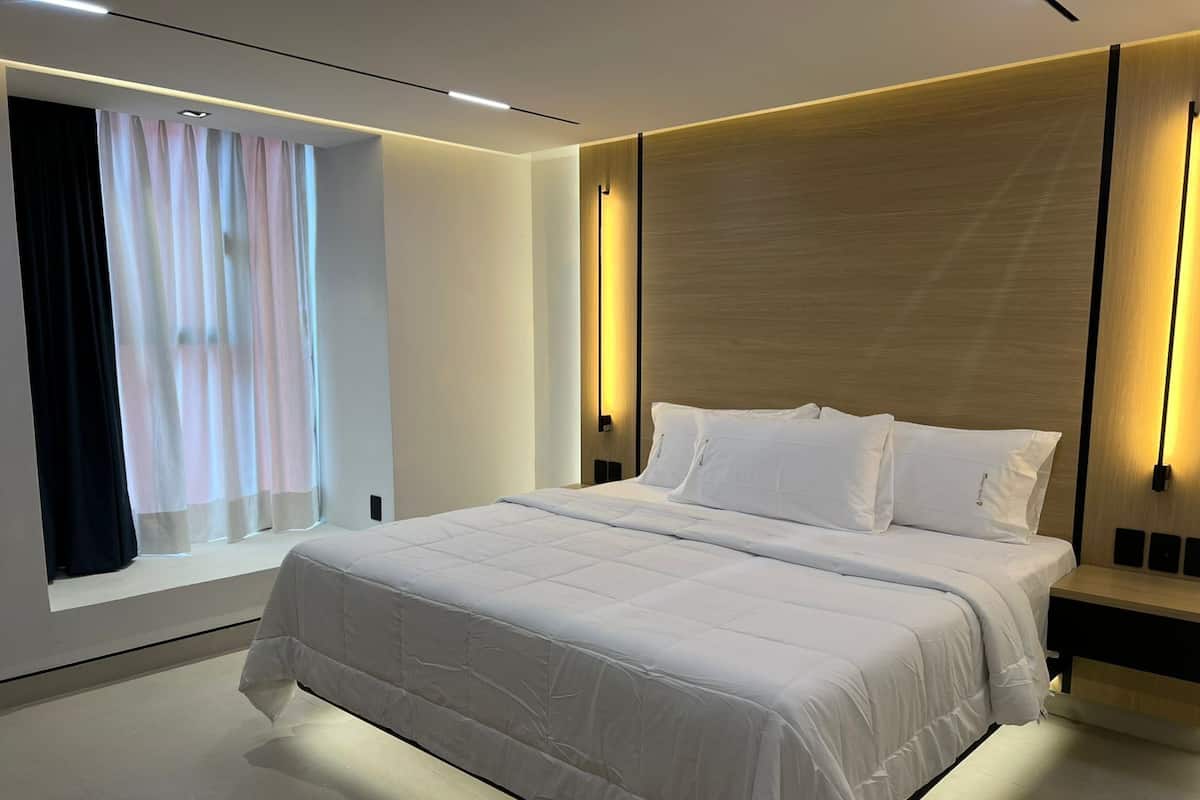 A modern bedroom with a large bed, a window with sheer curtains, and wall-mounted lighting.