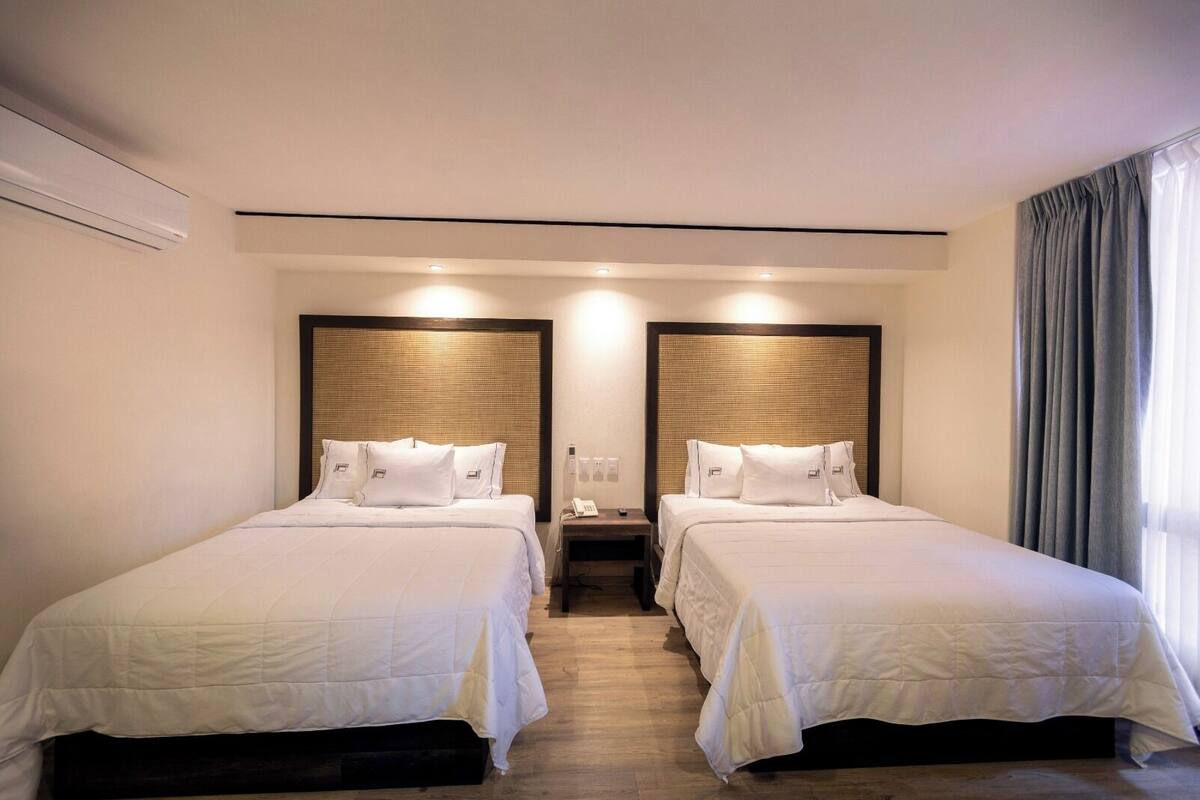A hotel room with two beds, each with white bedding and headboards. There is a nightstand between the beds with a phone and a lamp. The room has a wooden floor and a window with curtains.