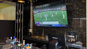 A bar area with a large screen displaying a football game, a counter with various bottles, and a coffee machine. - Hotel Lepanto Reforma (Mexico City)