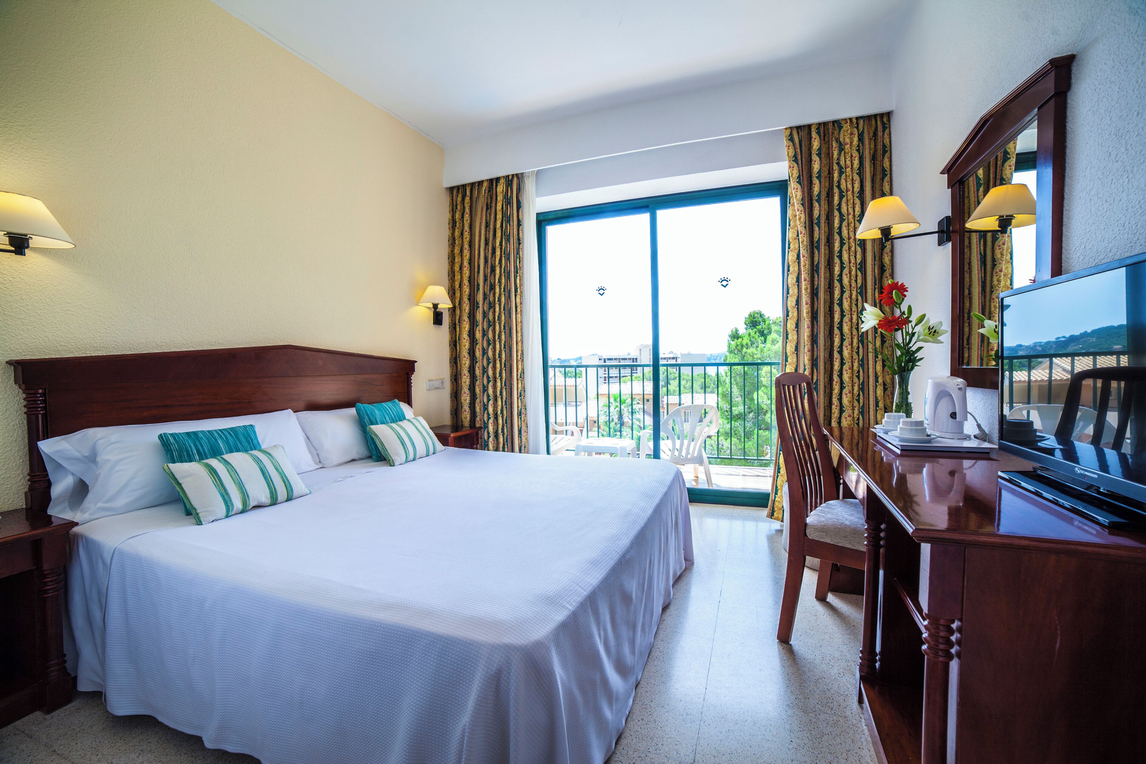 A hotel room with a large bed, a desk with a television, a chair, and a balcony with curtains.