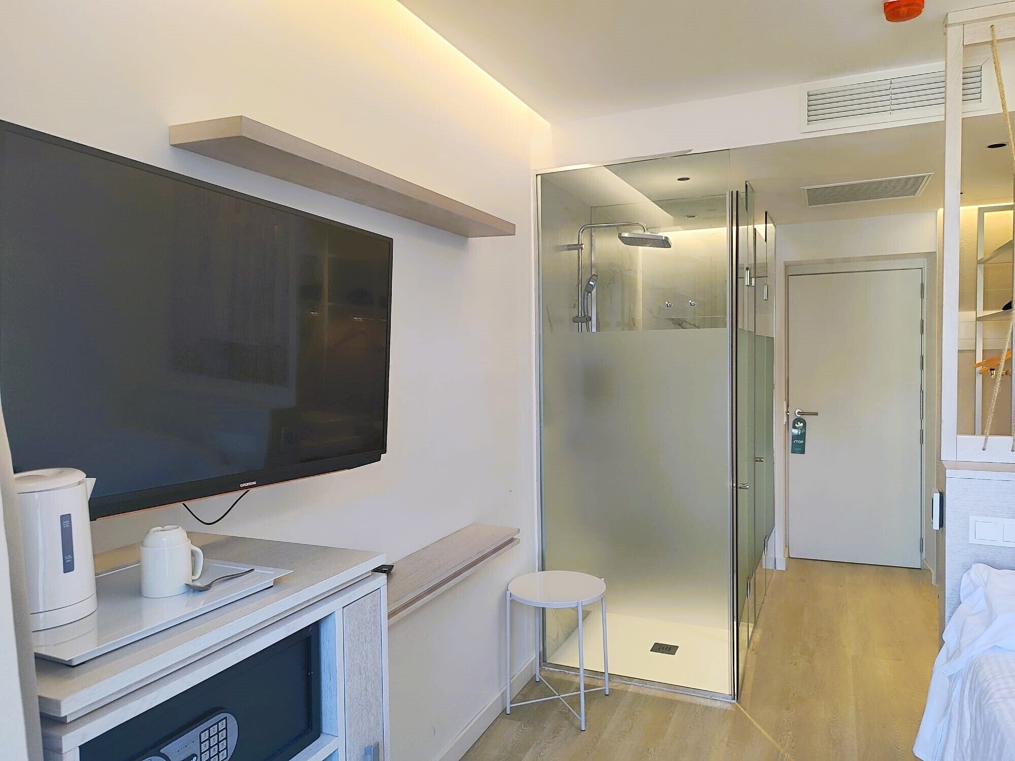 A modern hotel room with a flat-screen TV, a coffee maker, a small stool, a glass-enclosed shower, and a bed with white bedding.
