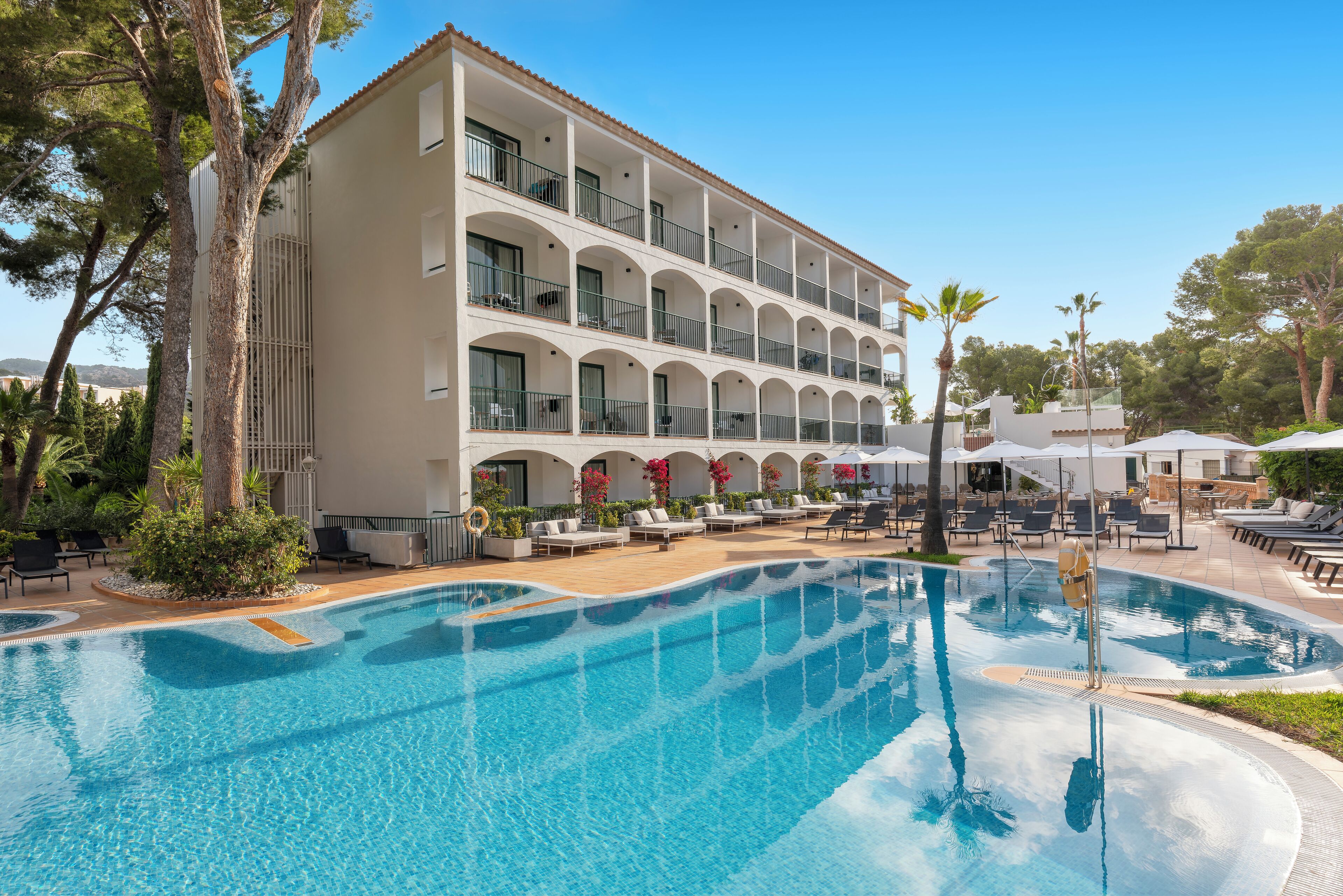 Un hotel con una piscina grande, mobiliario exterior y un edificio de varios pisos con balcones.