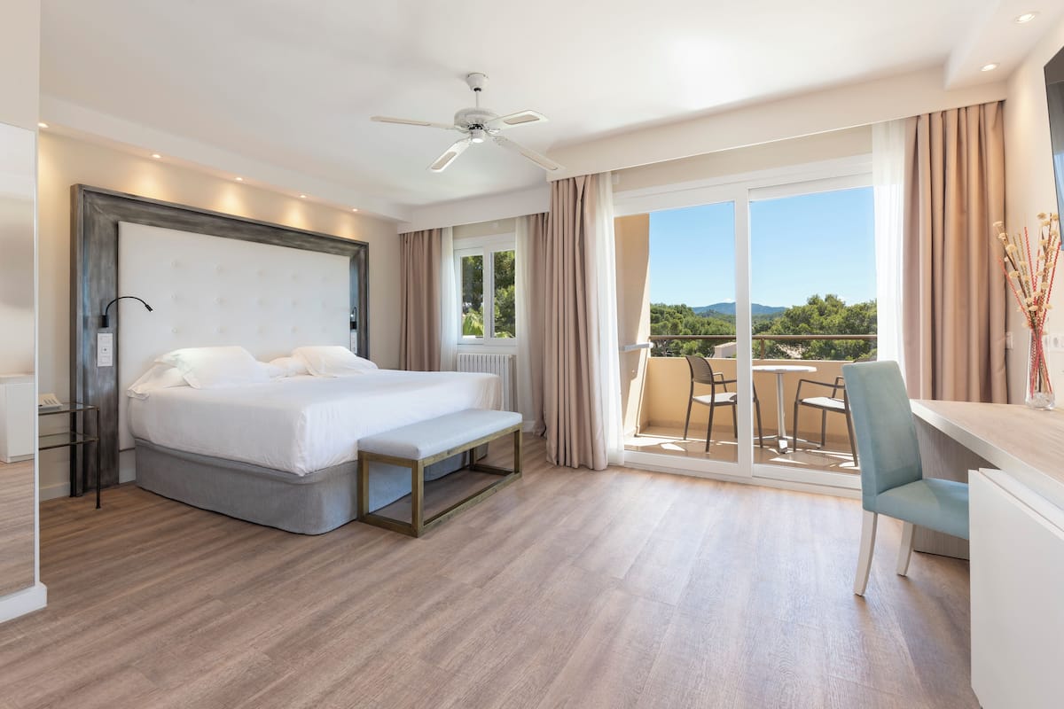 A modern hotel room with a large bed, a desk, and a balcony with a view of the landscape.